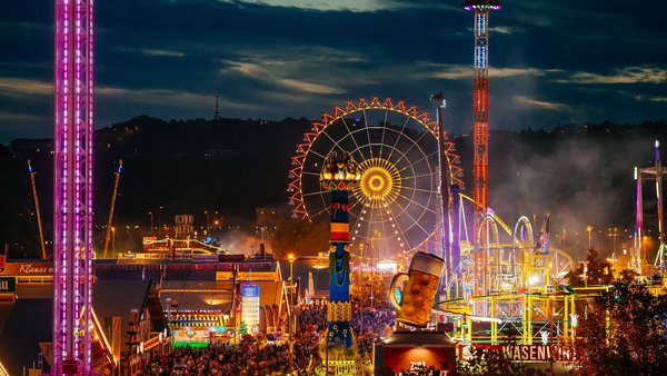 Blick auf den Festplatz bei Nacht mit den Lichtern der vielen Fahrgeschäfte. Dominant das beleuchtete Riesenrad, die Fruchtsäule und das Riesenbier auf dem Festzelt Wasenwirt.