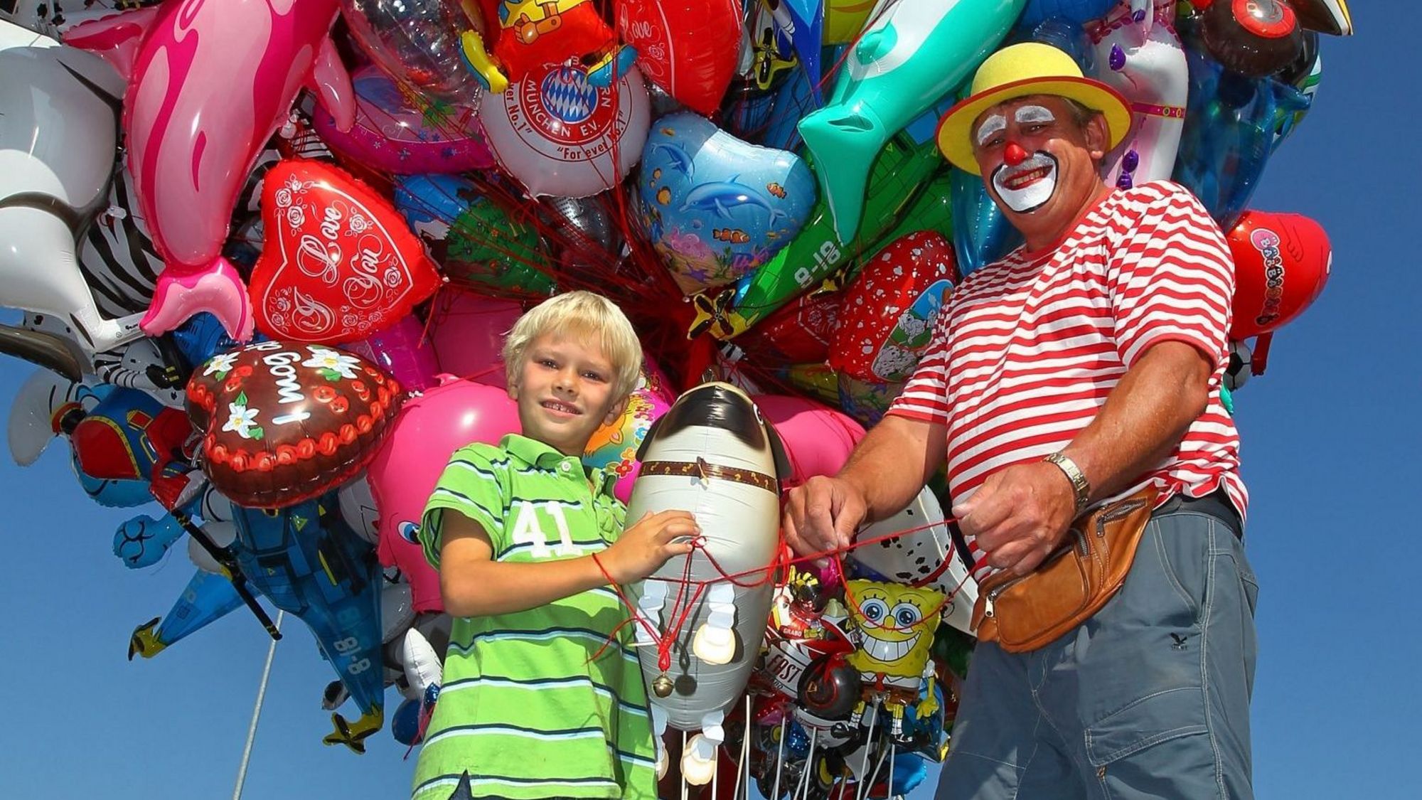 Ein Luftballon-Stand mit zahlreichen Luftballons, ein Junge kauft sich gerade einen Ballon beim Verkäufer und blickt in die Kamera