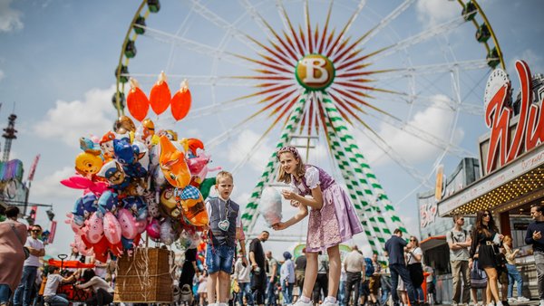 [Translate to English:] Zwei Kinder stehen vor dem Riesenrad und essen Zuckerwatte. Links von ihnen sehr viele bunte Ballons.