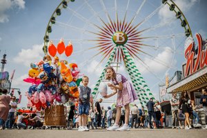 [Translate to English:] Zwei Kinder stehen vor dem Riesenrad und essen Zuckerwatte. Links von ihnen sehr viele bunte Ballons.