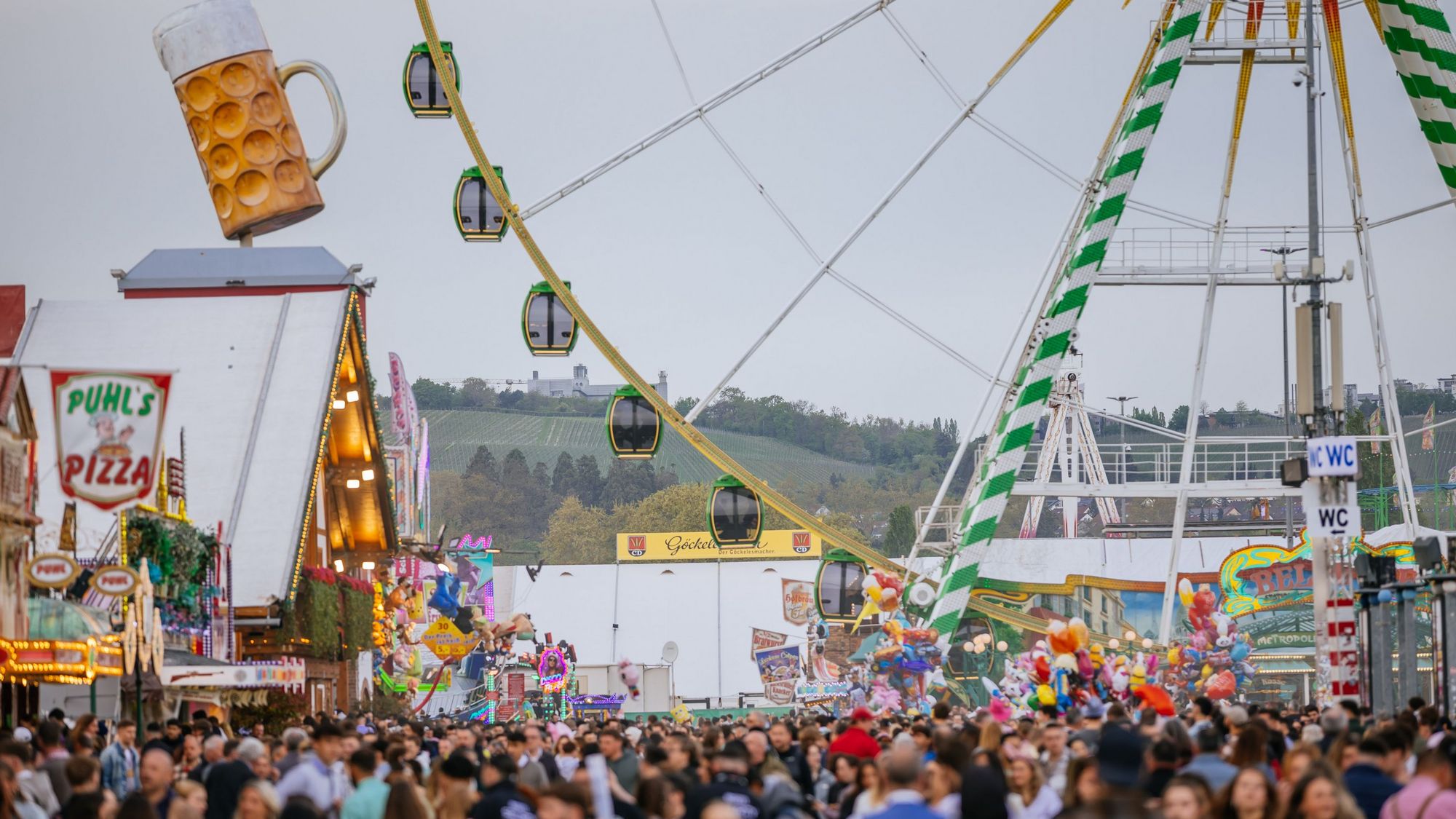 Eine Menschenmenge bahnt sich den Weg über den Cannstatter Wasen beim Frühlingsfest, vorbei an den Zelten. Im Hintergrund steht das Riesenrad